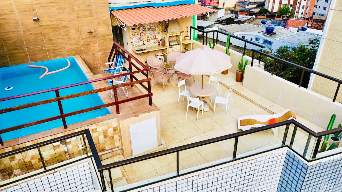 An outdoor space showcases a private pool alongside a designated area for dining. A round table with chairs is positioned beneath an umbrella, while a barbecue area is visible in the background. The surrounding walls are adorned with potted plants, complementing the urban backdrop.