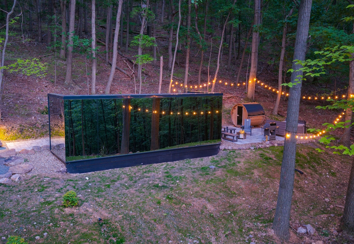 A modern mirrored villa is situated in a forested area, reflecting the natural surroundings. Soft, warm string lights are seen in the background, illuminating a cozy outdoor seating arrangement near a barrel sauna, enhancing the inviting outdoor space.
