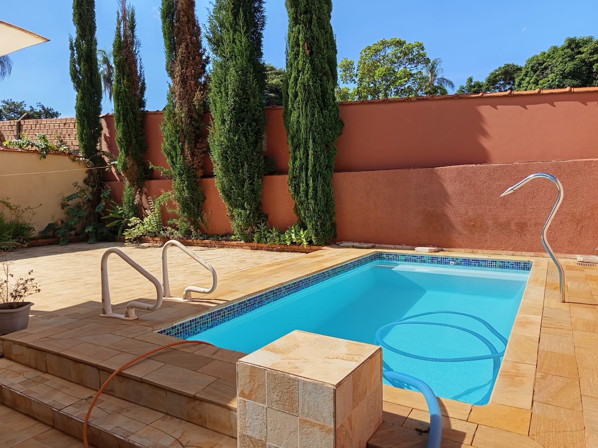 A rectangular swimming pool is surrounded by a stone patio, offering ample space for relaxation. Tall, green cypress trees line the back, providing privacy. A handrail and gentle steps lead into the pool, which is illuminated by sunlight on a clear day.