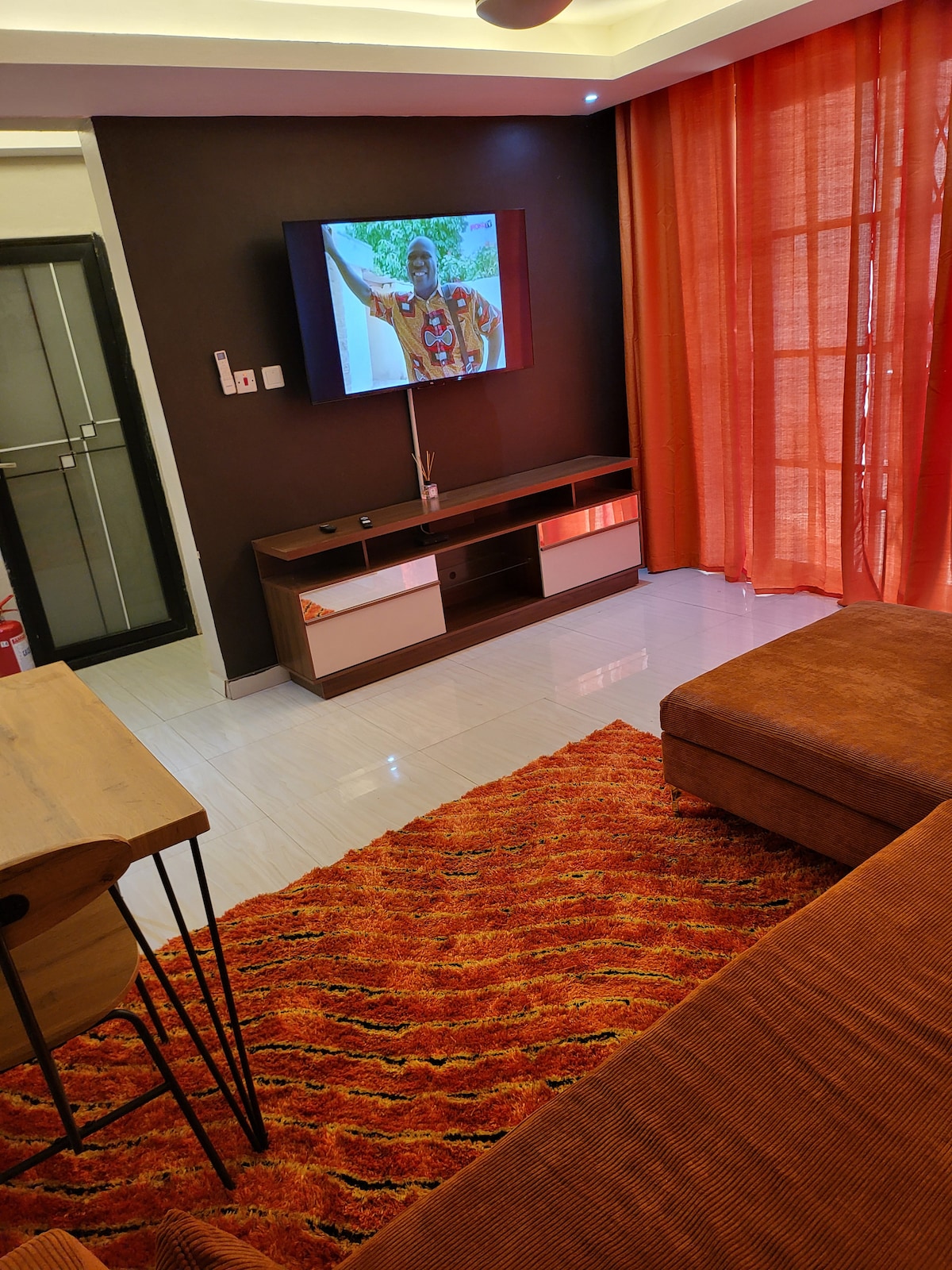 A modern living area features a large flat-screen television mounted on a dark wall above a stylish media console. A vibrant orange rug covers the floor, complementing the warm curtains that allow natural light to filter in, creating a cozy atmosphere.