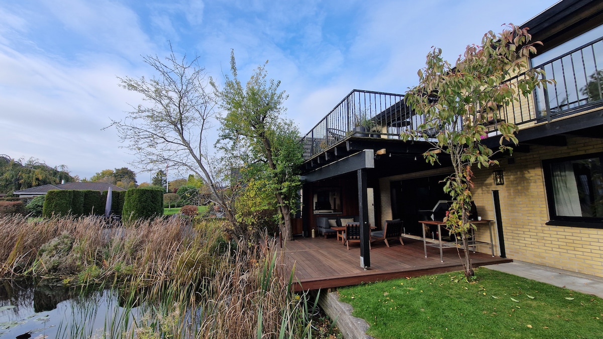 A wooden deck extends from the villa, furnished with outdoor seating. Lush greenery and tall grasses are seen surrounding the water's edge, while the upper terrace provides a view of the tranquil landscape. The building features large windows reflecting the daylight.