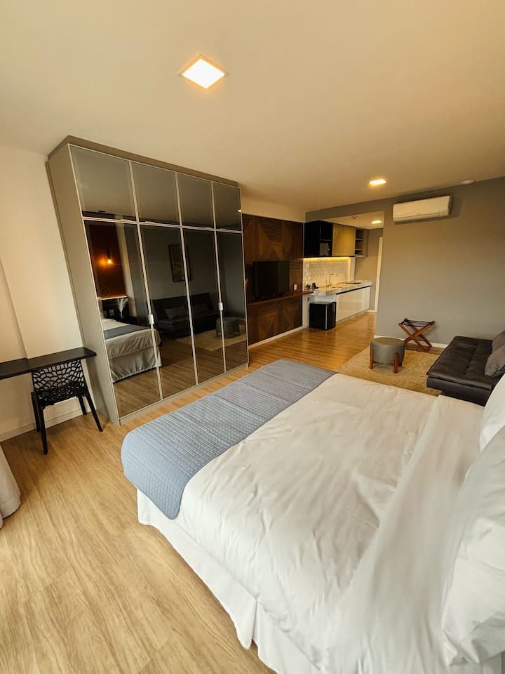 A well-organized bedroom area is visible, featuring a spacious bed with soft linens. A large mirrored wardrobe enhances the sense of space. A cozy seating area includes a dark sofa and a small table, with a kitchenette visible in the background, complemented by warm lighting.