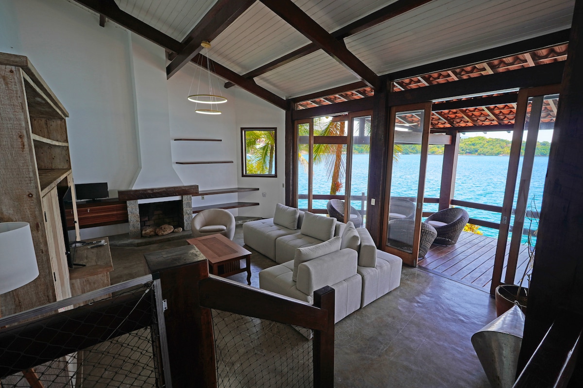 A bright living area features large windows overlooking the water, providing ample natural light. Comfortable, neutral-colored sofas are arranged around a small wooden table, while a fireplace adds a cozy touch. Decor elements include simple wall art and a pendant light, enhancing the warm ambiance.