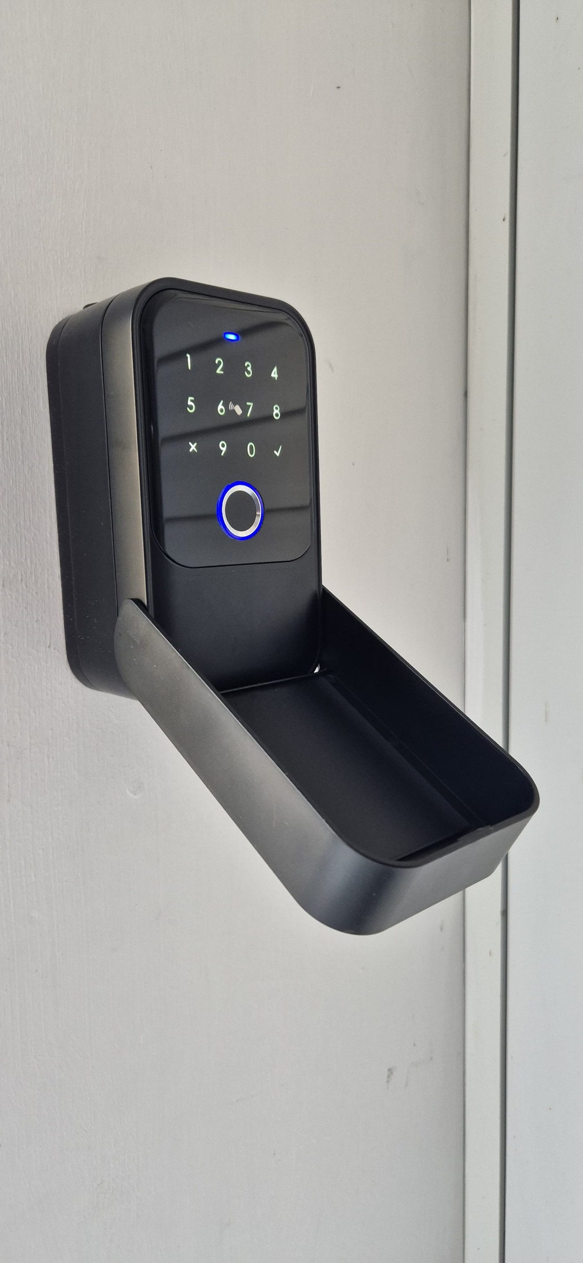 A digital keypad lock is mounted on a white door. The lock includes numerical buttons and a blue circular indicator light. Below the keypad, a shallow tray is designed to catch items. The overall finish appears sleek and modern.