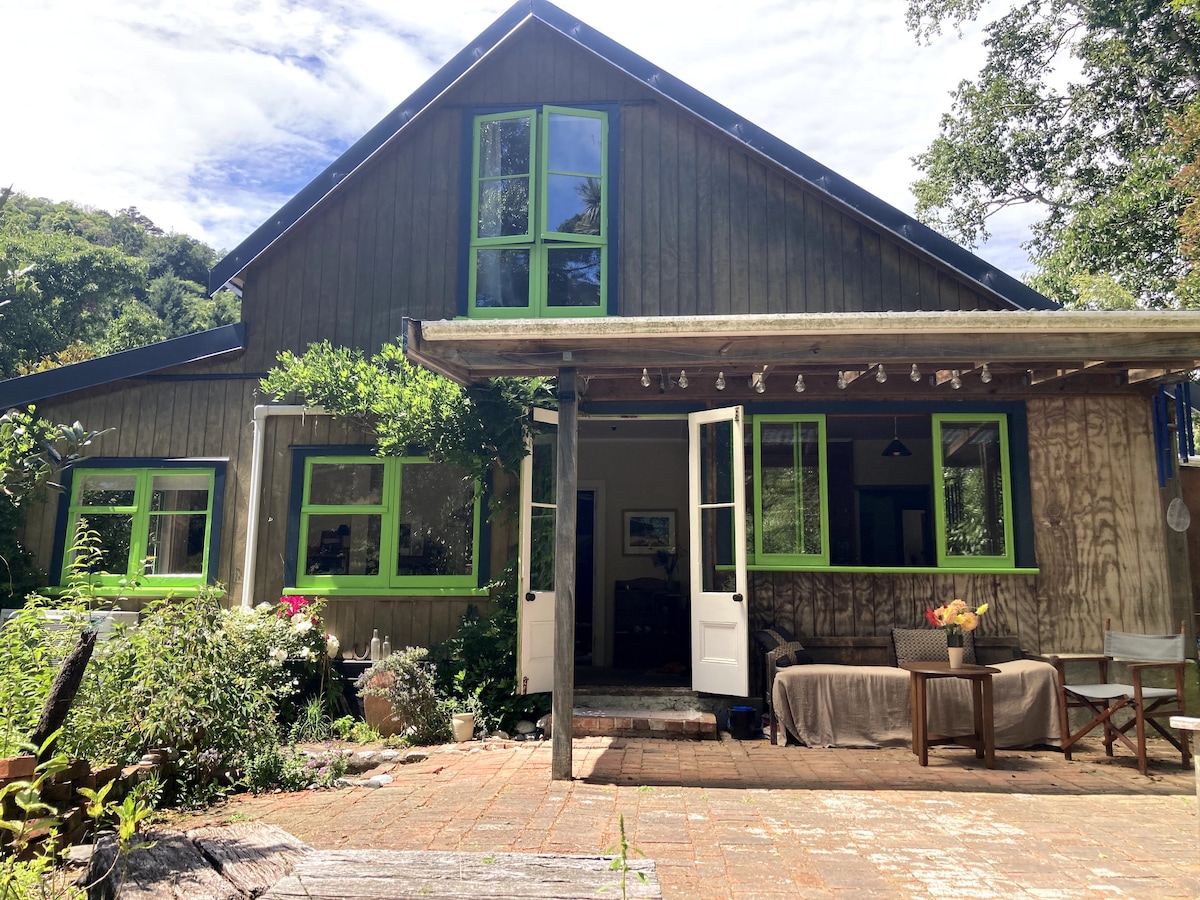 The exterior of the wooden house features green window frames and a welcoming porch area. A spacious outdoor patio is adorned with seating arrangements and flower pots, surrounded by lush greenery, inviting guests to enjoy the peaceful environment.