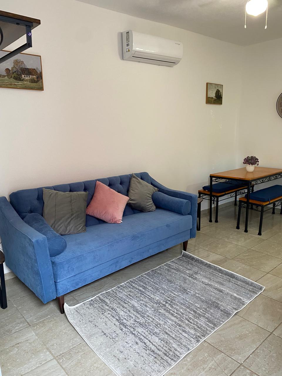 A blue upholstered sofa is positioned against a light-colored wall, accented by decorative throw pillows in soft hues. A small dining table with four chairs is visible, along with a rug placed beneath. Air conditioning is mounted above for climate control.