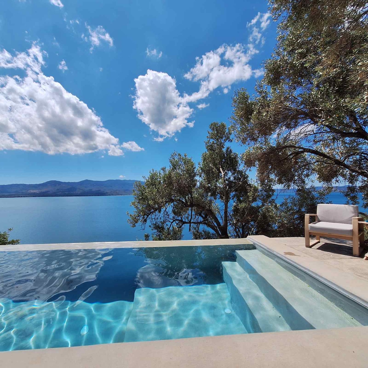 A private pool is visible, featuring clear, shimmering water that reflects the surrounding landscape. Trees provide shade in the area, and a scenic view of the bay stretches beyond, with blue skies and clouds enhancing the serene atmosphere.