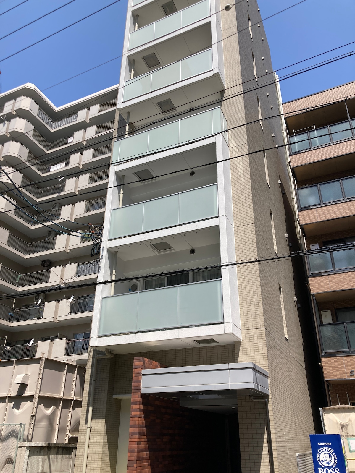 A modern apartment building is displayed, showcasing five stories with a minimalist facade. The structure features large glass balconies on each floor, providing a bright appearance. The buildings on either side illustrate a blend of architectural styles typical to urban areas.
