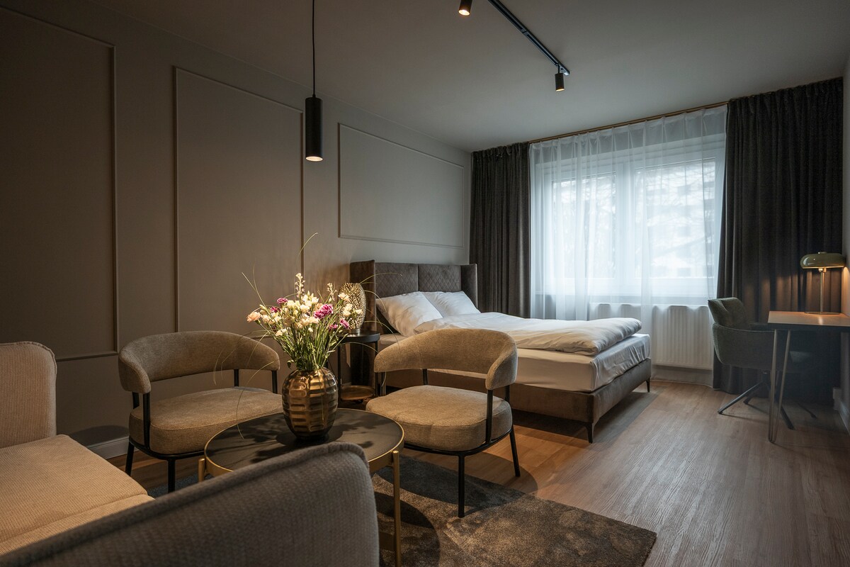 A serene room features a spacious bed with light bedding, complemented by two plush chairs and a round table. Natural light filters through a large window, and a work desk is situated near the curtains, enhancing the room's functionality.