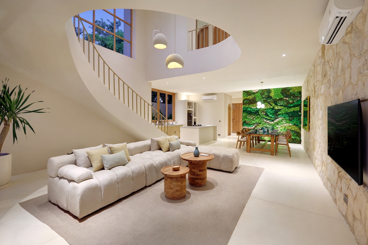An open-plan living area features a spacious sofa with light-colored upholstery and two wooden coffee tables. A soft area rug anchors the seating, while a curved staircase leads to the upper level. A green wall art piece adds a touch of nature, and large windows allow natural light in.