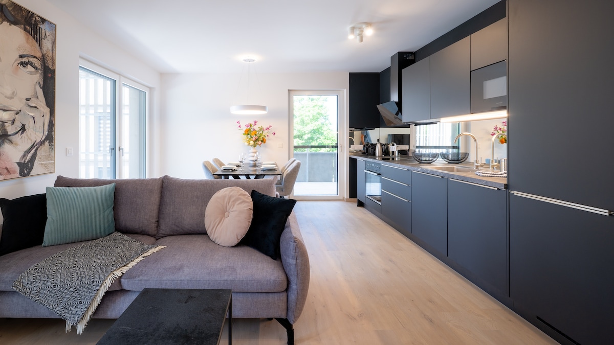A contemporary living space features an open layout with sleek cabinetry and a modern couch. A dining area is visible in the background, with a table set for several guests. Natural light fills the room through large windows, enhancing the neutral tones of the decor.