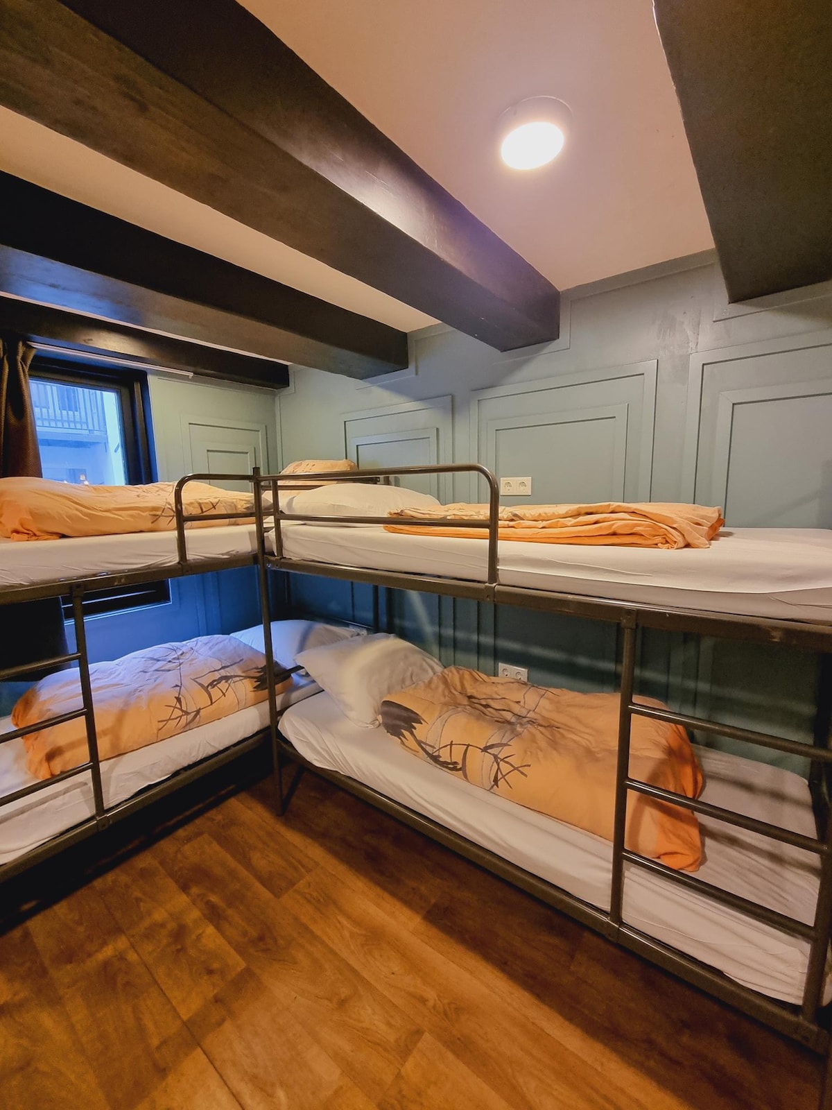A dormitory room is presented with two sets of sturdy bunk beds, each with neatly arranged bedding featuring an earthy design. Natural light enters through a window, illuminating the wooden floor and muted green walls, creating a cozy atmosphere.