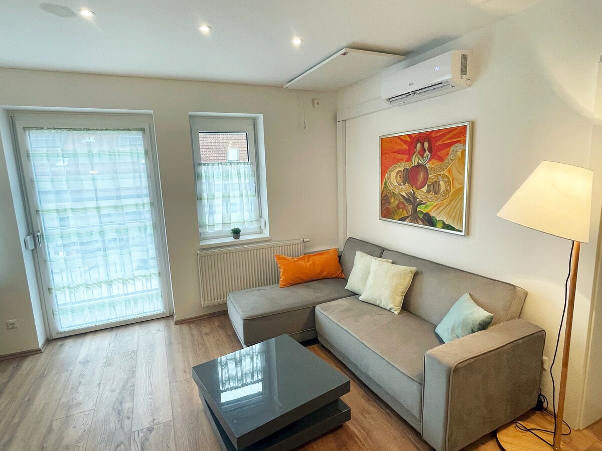A well-lit living area features a comfortable sectional sofa with decorative cushions. A coffee table sits prominently in front, while large windows with sheer curtains allow natural light to fill the space. An artwork adorns the wall, adding a splash of color.