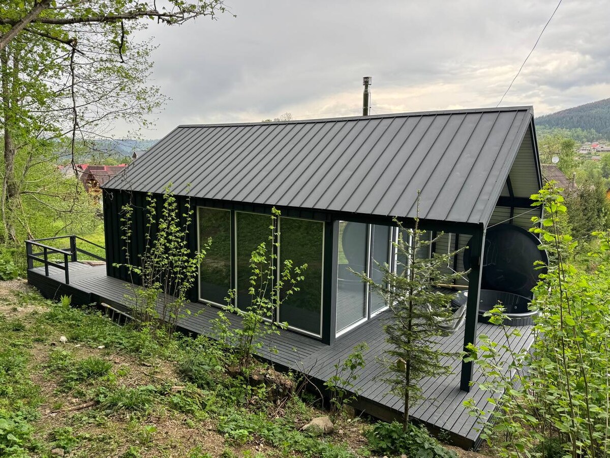 The modular house is set amidst greenery, featuring a sloped black roof and large glass panels. A wooden deck extends from the structure, offering space to enjoy the natural surroundings. Vegetation and trees frame the scene, creating a serene atmosphere.