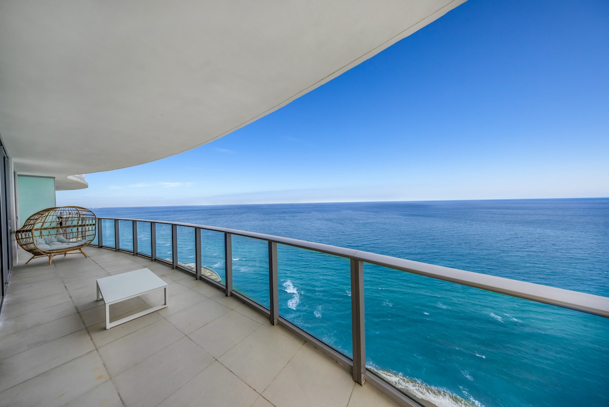 A spacious private balcony is showcased, featuring a comfortable seating area and a low table. The expansive ocean view is visible, with clear blue skies and gentle waves extending toward the horizon, creating a serene and tranquil environment.
