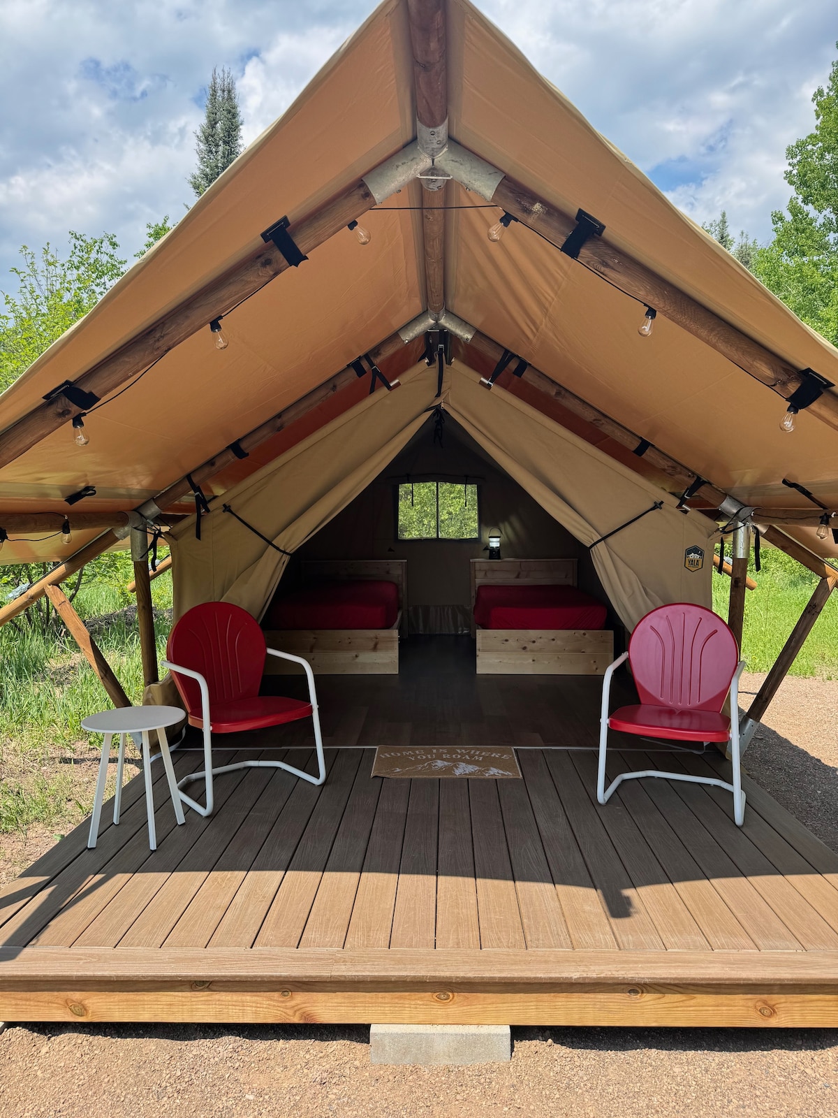 A glamping tent features a spacious deck, inviting two red chairs and a small table. Inside, two beds with wooden frames are visible. A window at the back allows natural light to enter, surrounded by greenery.