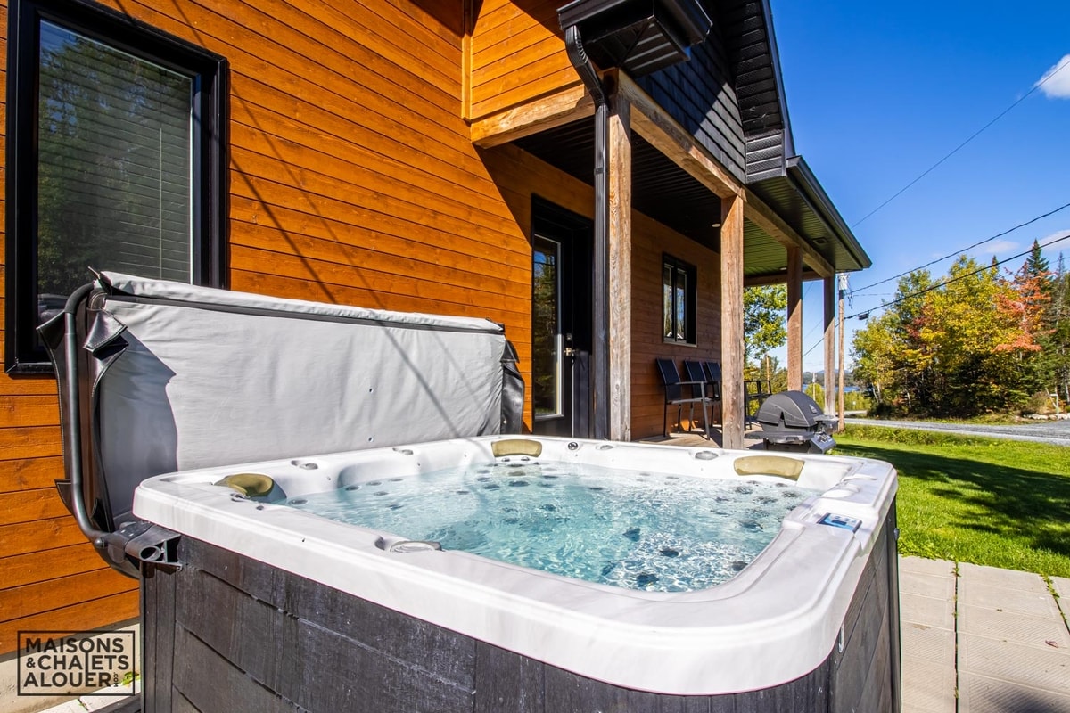 A private hot tub is positioned next to the chalet, featuring bubbles rising from the water's surface. The wooden exterior of the chalet is visible in the background, complemented by a well-maintained lawn and outdoor seating.
