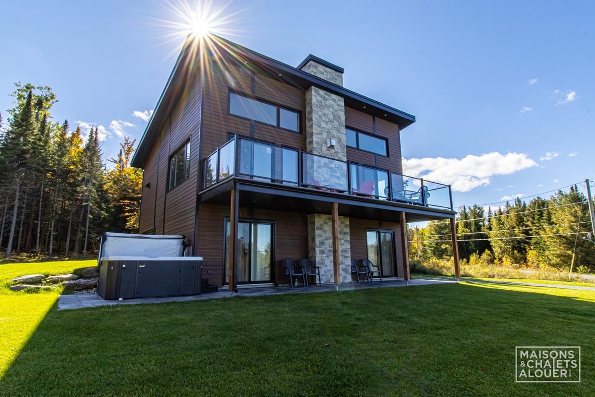 The exterior of a modern chalet is displayed, featuring a two-story structure with large windows and a stone accent wall. A spacious balcony is visible on the second floor, overlooking a well-maintained lawn. The property is surrounded by trees, enhancing its natural setting.