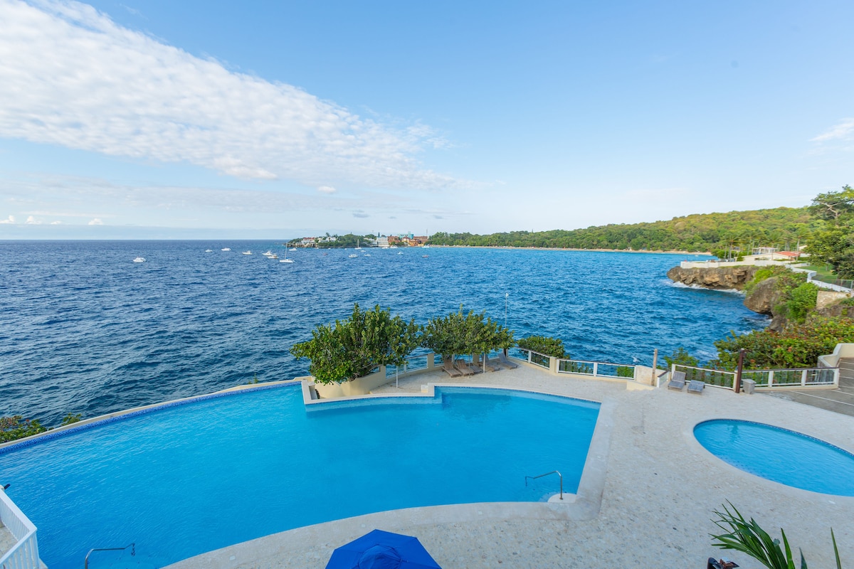 An expansive outdoor pool is surrounded by lush greenery and offers ocean views. The clear blue water reflects the sky, and the pool features a gentle gradient. Nearby, a terrace can be seen, providing additional space for relaxation and enjoying the scenery.