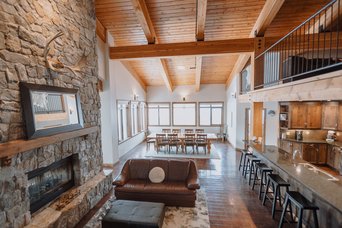 A spacious living area features a stone fireplace complemented by wooden beams and large windows allowing natural light. A plush sofa sits on an area rug, while a dining table accommodates multiple guests. The kitchen bar with stools is visible in the background.