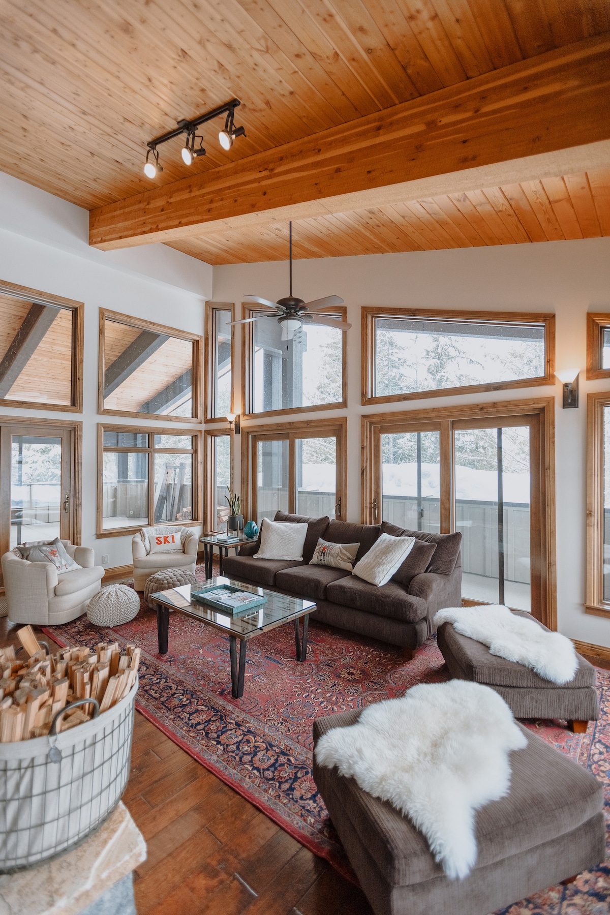 A spacious living area features vaulted wooden ceilings and large windows, allowing natural light to fill the room. Comfortable seating is arranged around a glass coffee table, with a patterned area rug beneath. A cozy chair and a decorative basket of firewood complete the inviting space.