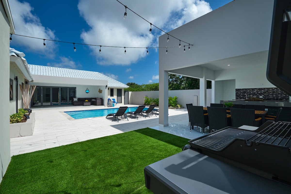 A private outdoor area features a sparkling pool surrounded by lounge chairs, with an outdoor kitchen and dining space nearby. String lights are suspended above the area, illuminating the serene setting. Lush green grass enhances the tropical ambiance.