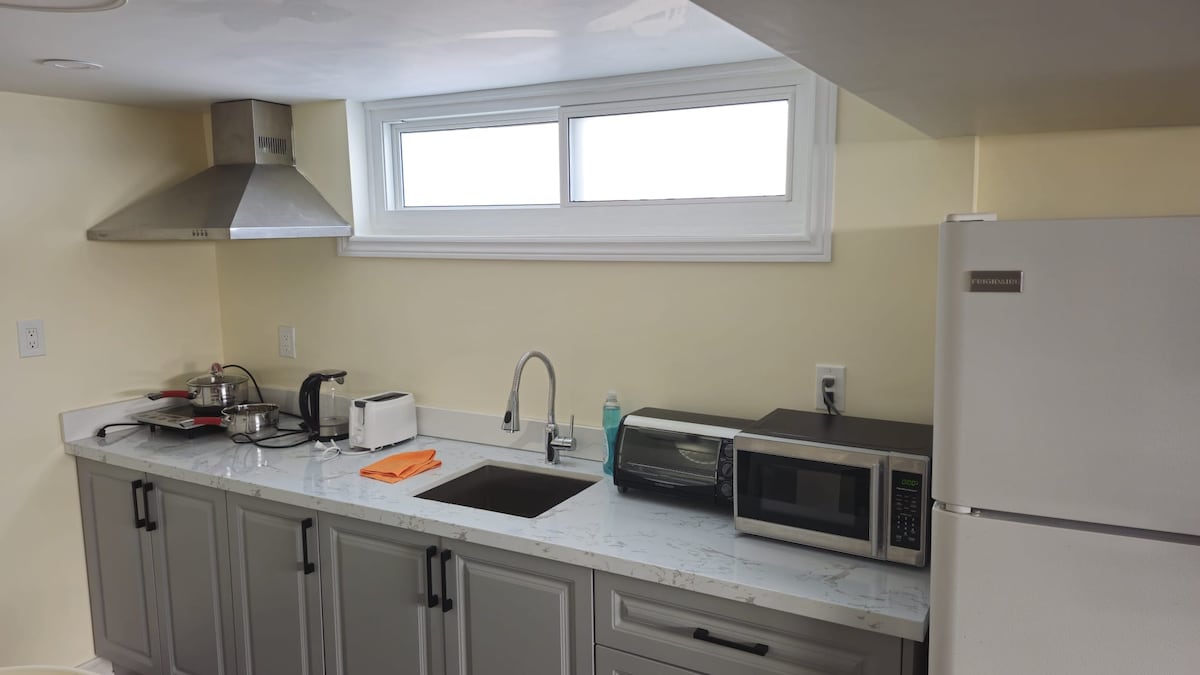 The kitchen features a light-colored countertop with ample workspace. A stainless-steel sink is positioned between a microwave and a refrigerator. Above, a ventilation hood is visible, and a window allows natural light to enter, creating a bright and practical cooking area.