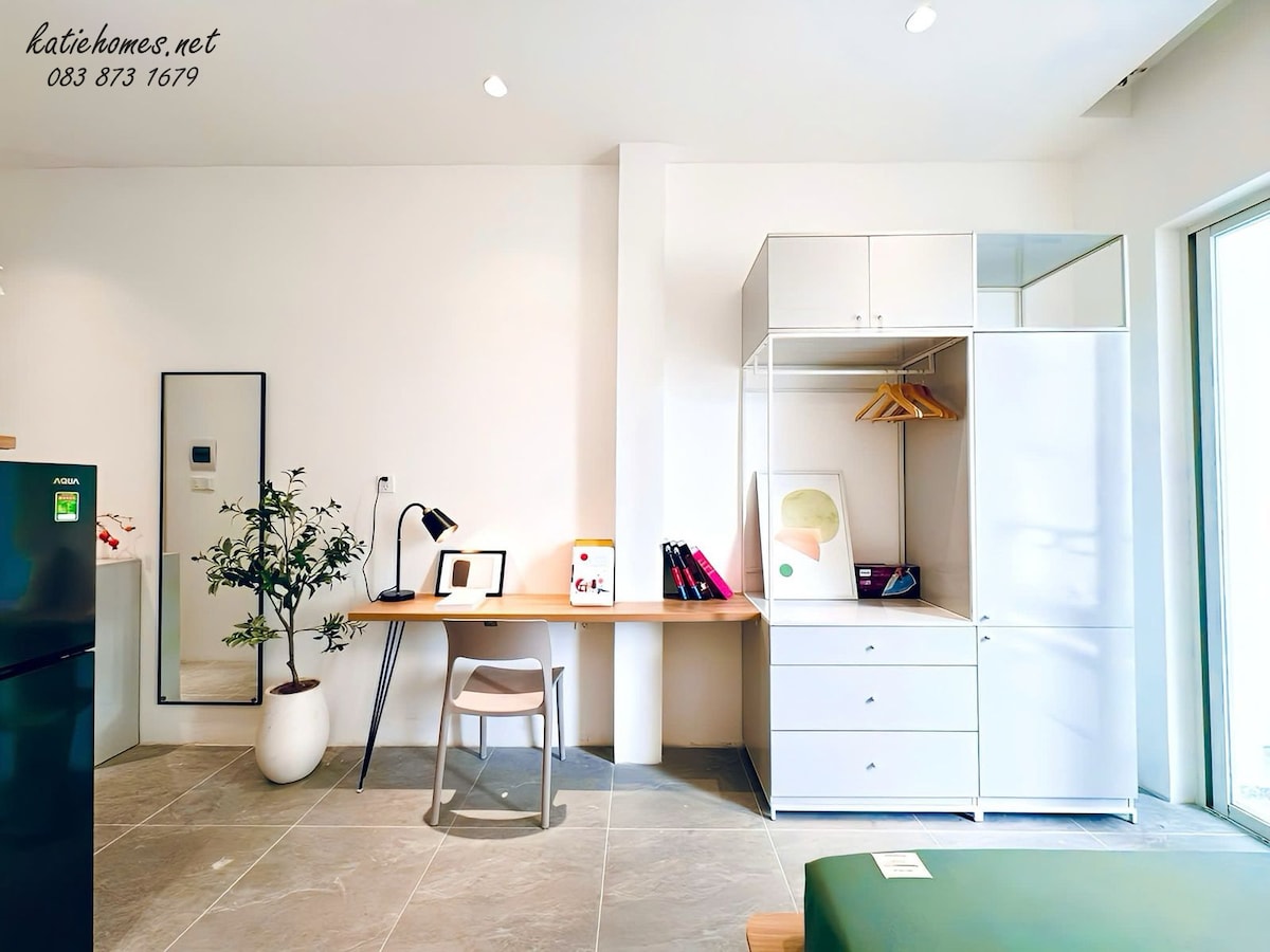 A bright and airy interior is highlighted by a wooden desk paired with a simple chair. A wardrobe and shelf contain neatly arranged items, while a small indoor plant adds a touch of greenery. Natural light streams through large windows, enhancing the spacious feel.