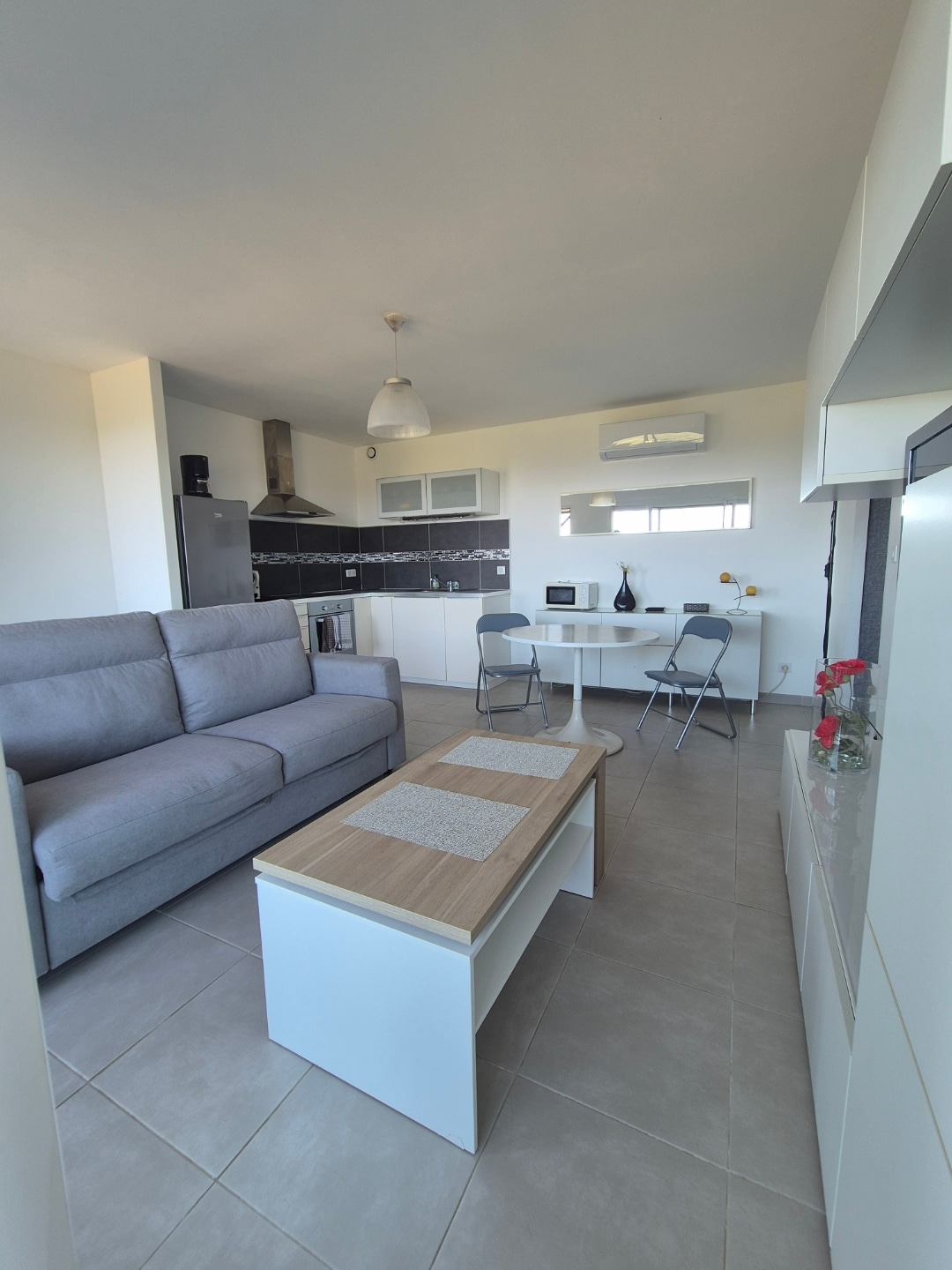 A well-organized living area features a light gray sofa facing a modern dining table with four chairs. A compact coffee table sits in front, complemented by decorative flowers. The kitchenette is visible in the background, complete with essential appliances and sleek cabinetry.