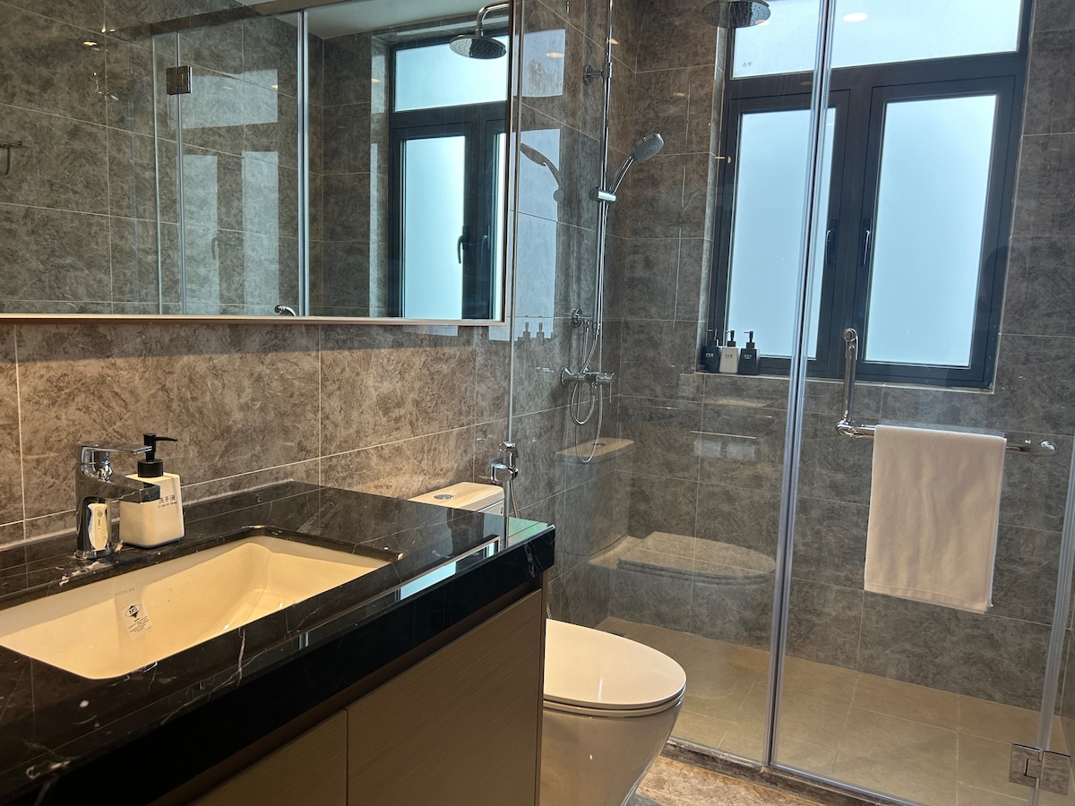 A modern bathroom is displayed, featuring a sleek vanity with a dark countertop and a large mirror. A glass-enclosed shower is visible, equipped with dual shower heads. Natural light filters through the frosted windows, enhancing the clean, contemporary design.