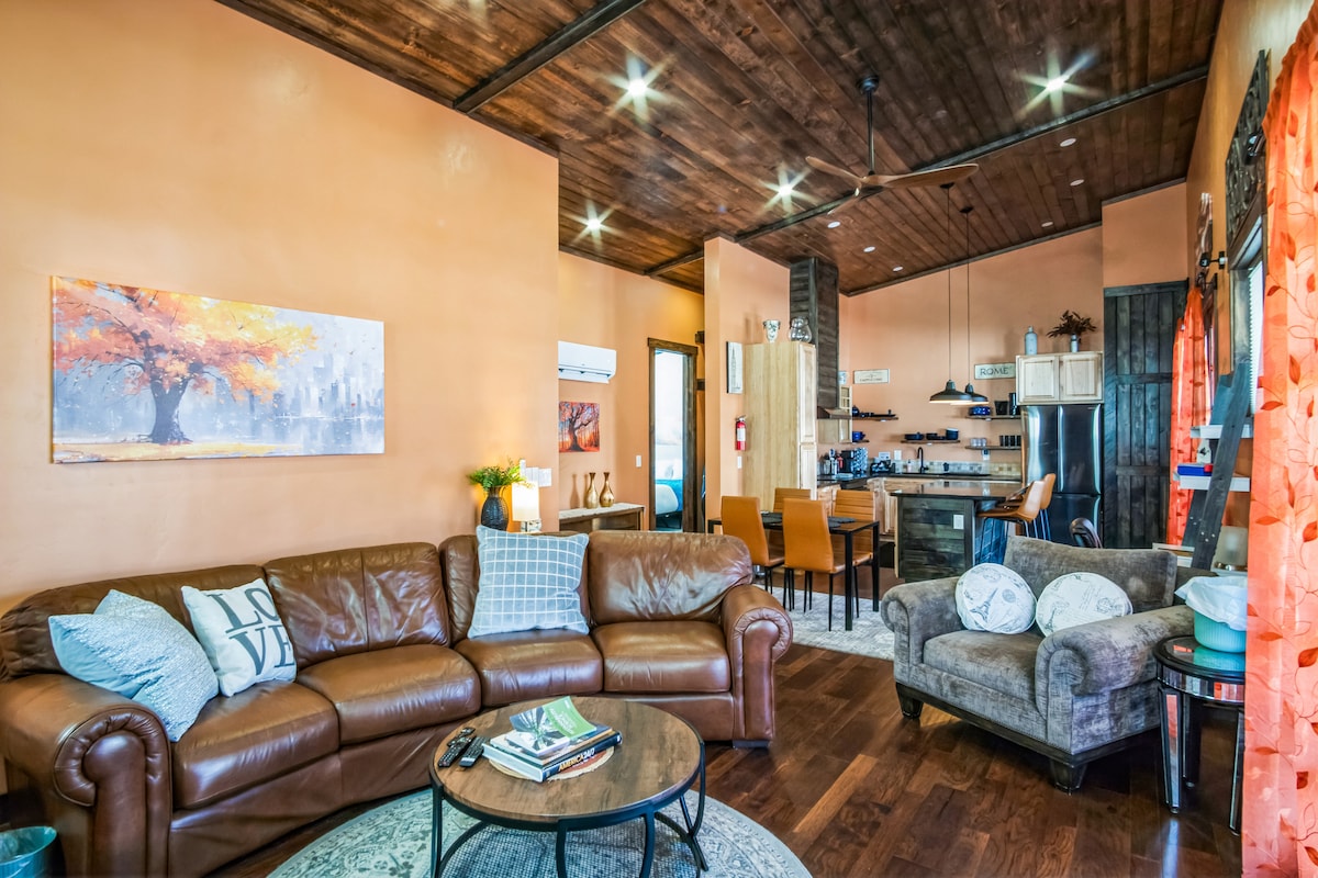 A spacious living area features a comfortable leather sofa paired with a cozy armchair. Tall ceilings with wooden beams add warmth, while natural light filters through bright orange curtains. A round coffee table occupies the center, surrounded by an inviting atmosphere leading to the dining area.