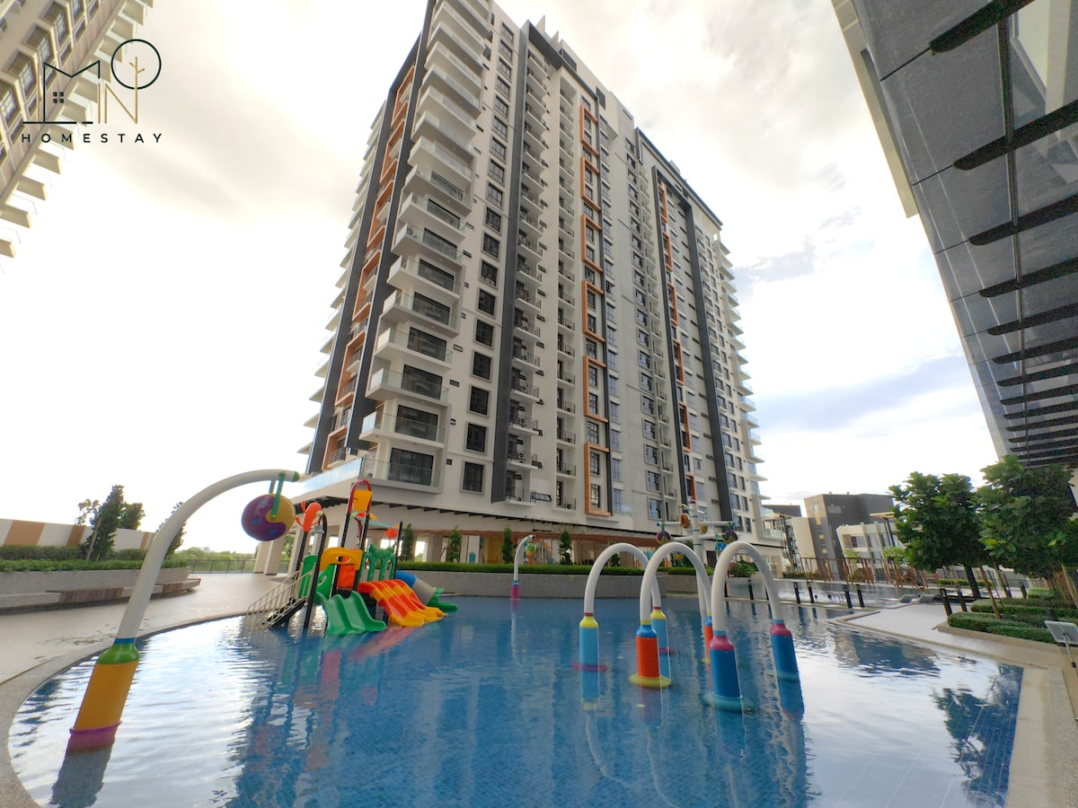 A modern multi-story building is depicted alongside a vibrant water play area, featuring colorful structures for children's recreation. The pool reflects the sky and surrounding greenery, creating a serene outdoor space for relaxation. Large windows on the building provide glimpses into the interior.