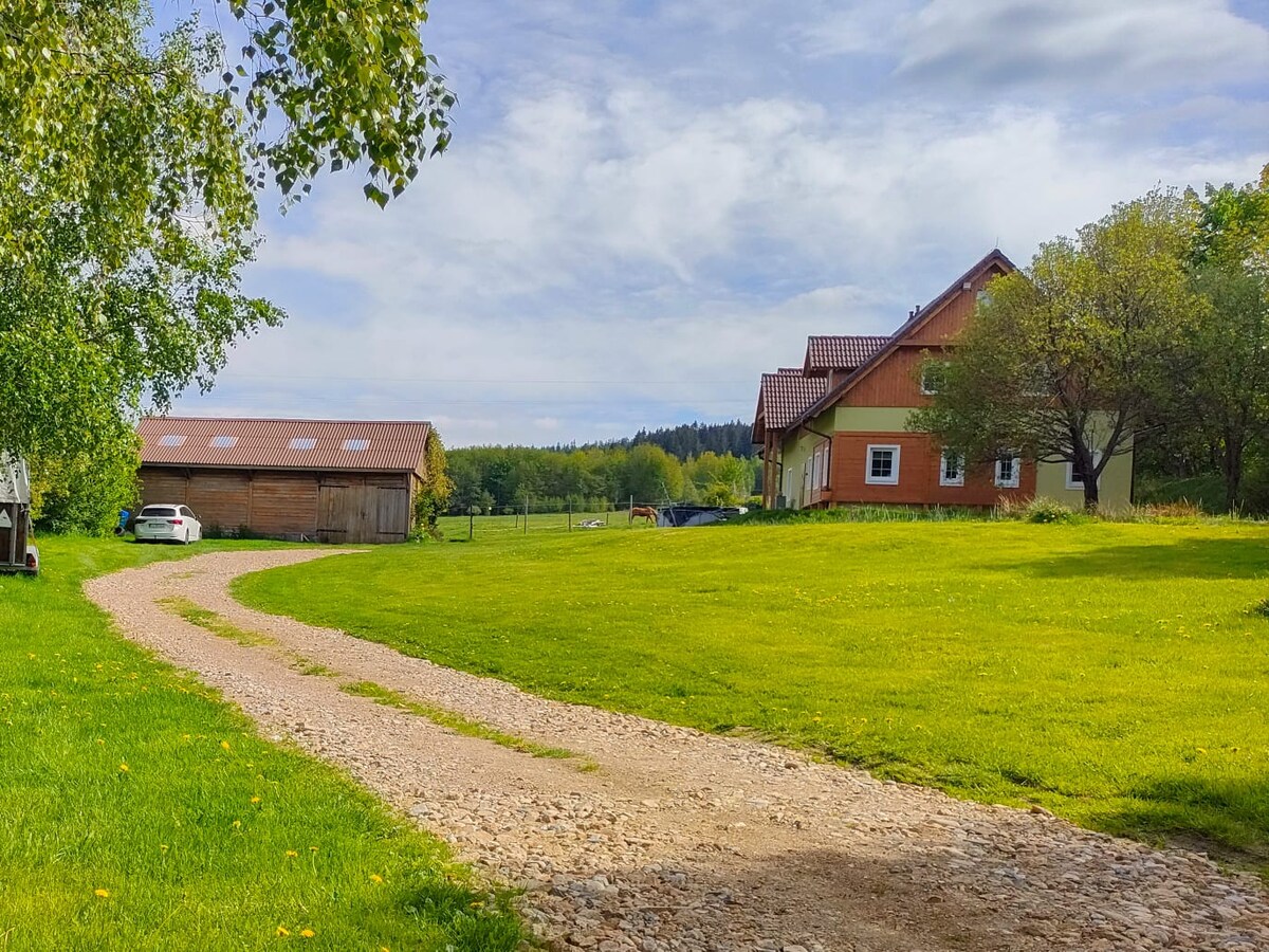 A winding gravel path leads through a lush green lawn towards a charming two-story house. The property is surrounded by trees and features a barn in the background, with rolling hills visible under a partly cloudy sky.