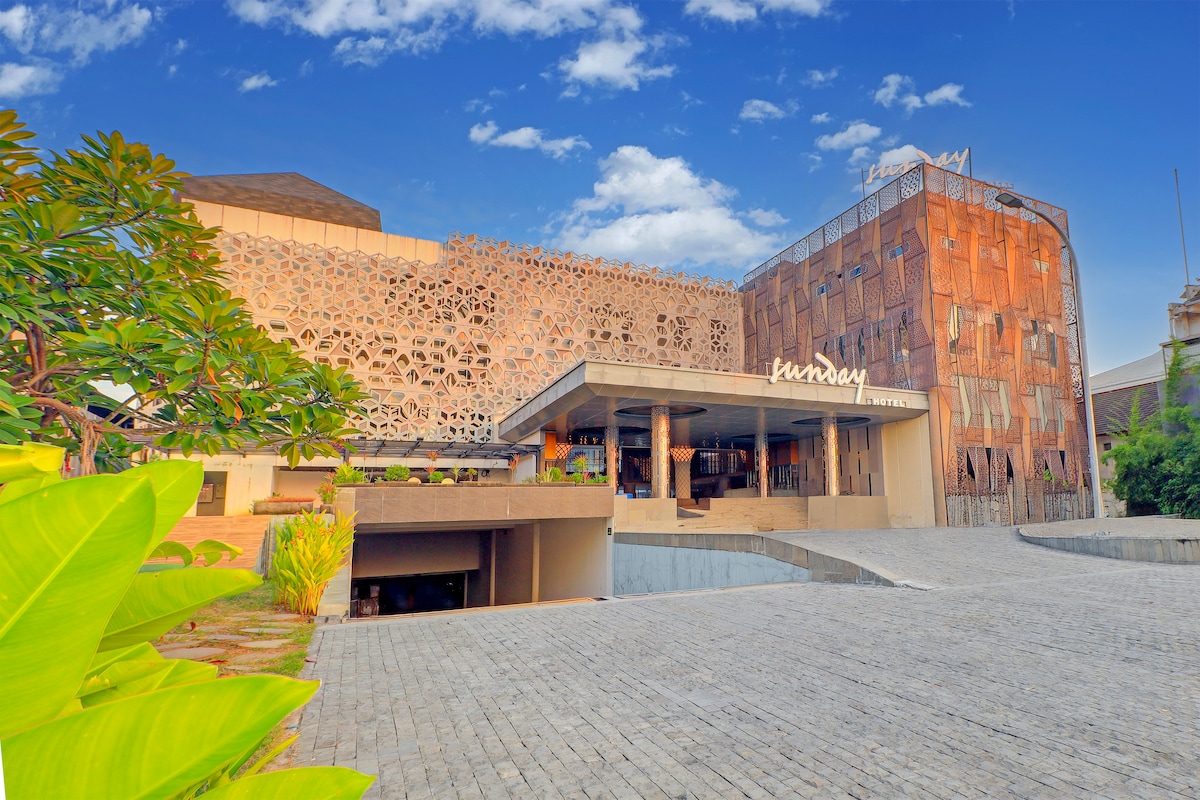 The exterior of Sunday Hotel Arshika showcases a blend of modern architecture and natural elements. A detailed patterned wall enhances the facade, while lush greenery and a paved pathway lead to the entrance. The blue sky adds to the inviting atmosphere of the property.
