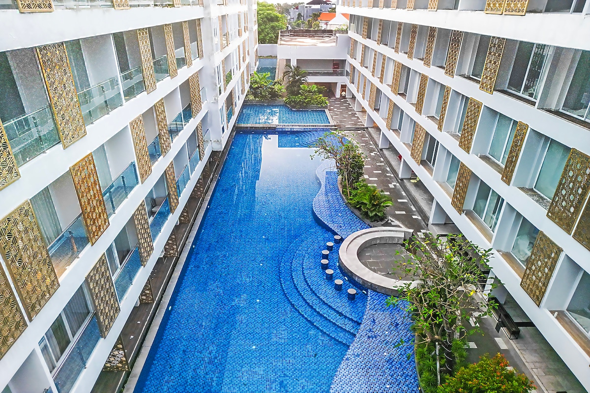 An aerial view captures a serene outdoor pool surrounded by modern architecture. The pool features a fluid design with steps leading into the water, complemented by meticulously landscaped greenery and lounge areas with seating around the perimeter.