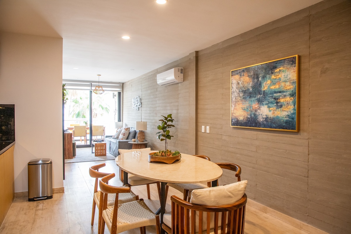 A dining area features a round marble table surrounded by six wooden chairs. A piece of wall art adds color to the textured wall. In the background, a living space is visible, with seating and natural light filtering through the glass doors that lead to an outdoor area.