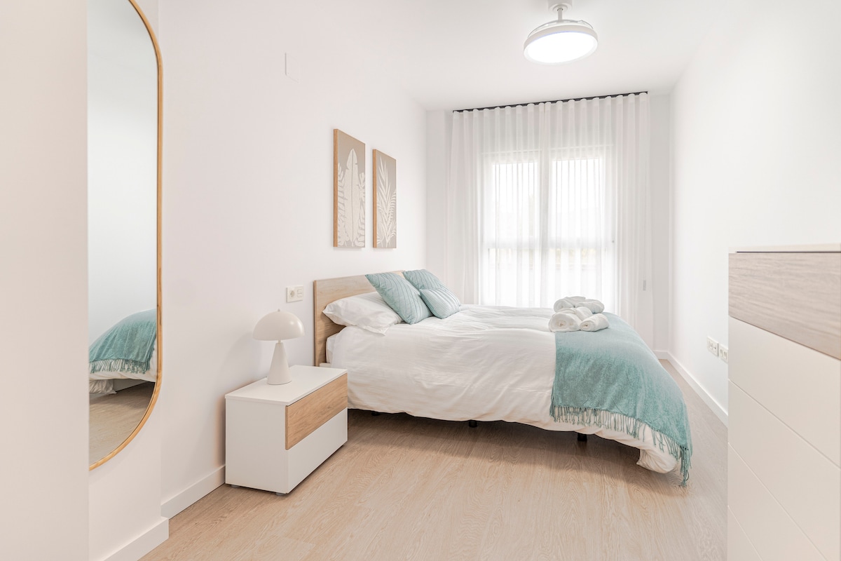 غرفة نوم بسرير مزدوج، وبياضات سرير بيضاء وبطانية فيروزية، ووسائد مطابقة، وطاولة بجانب السرير مع مصباح، وخزانة، وخزانة، ومرآة عمودية، ولوحتين زخرفيتين ونافذة مع ستائر شفافة، مع وصول مباشر إلى الحمام.
