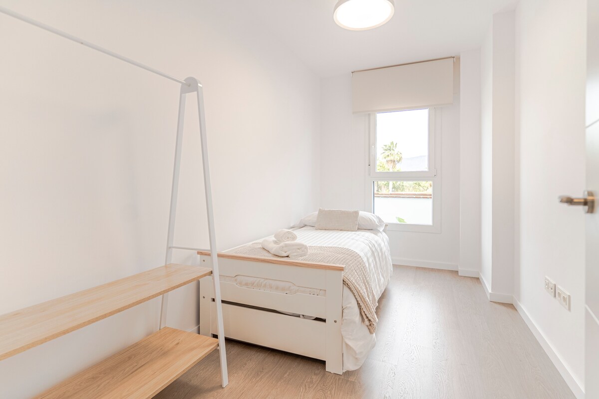 غرفة نوم مع سرير فردي، وبياضات بيضاء ومناشف، ورف معلق مفتوح مع أرفف وشماعات، وأرضية خشبية فاتحة، ونافذة مع ستارة دوارة وإمكانية الوصول المباشر إلى الردهة ومنطقة الحمام، في مساحة صغيرة وعملية.
