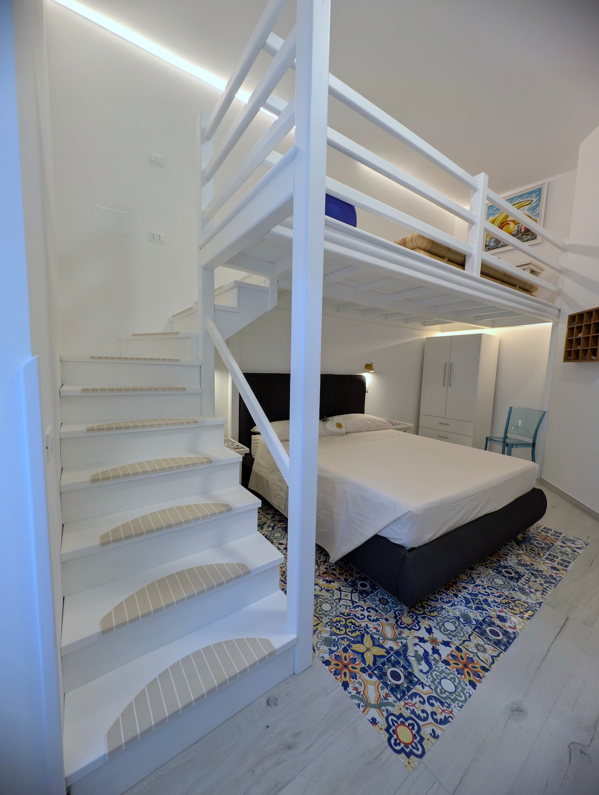 A lofted sleeping area is positioned above a spacious bed, providing vertical depth to the room. Light wood stairs lead to the upper level. Neutral tones complement colorful patterned tiles on the floor, creating a vibrant yet serene space. Ample natural light fills the area.