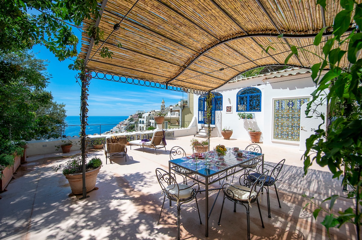 An outdoor terrace features a large dining table surrounded by elegant chairs. The area is shaded by a woven canopy, with terracotta pots displaying vibrant greenery. Scenic sea views and charming coastal architecture are visible in the background, creating a serene dining atmosphere.