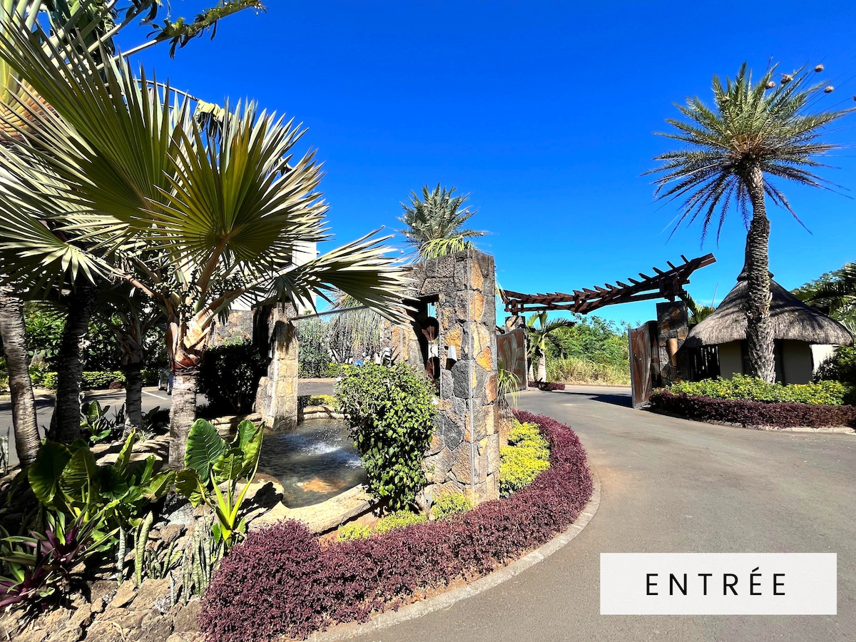 The entrance to the residence features neatly arranged palm trees and vibrant foliage, framing a stone path that leads into the property. A clear blue sky is visible above, creating a bright and welcoming ambiance.