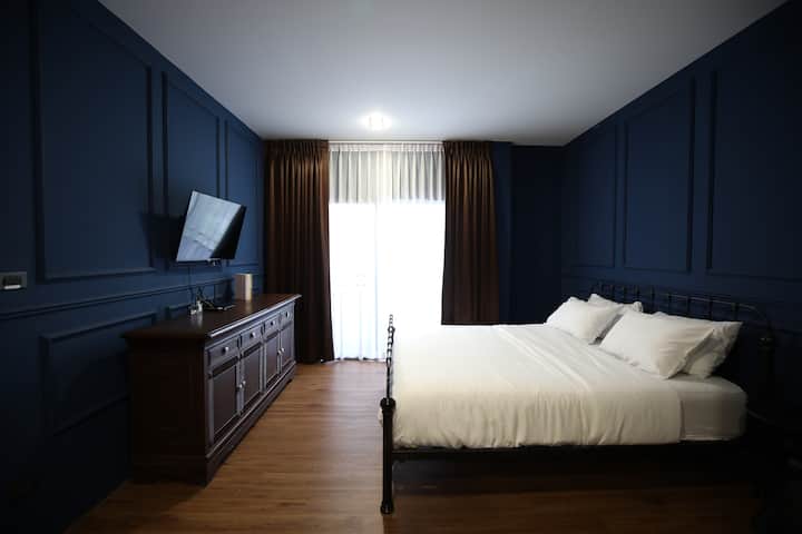 Chumba cha kulala cha kustaajabisha, cha kifahari kilicho na kuta za rangi ya bluu, fremu nyeusi ya chuma, mashuka meupe, na mapazia yenye rangi ya hudhurungi. Chumba hicho kina kitambaa cha mbao cha zamani kilicho na televisheni iliyowekwa, inayofaa kwa ajili ya kupumzika kwa mtindo usio na wakati