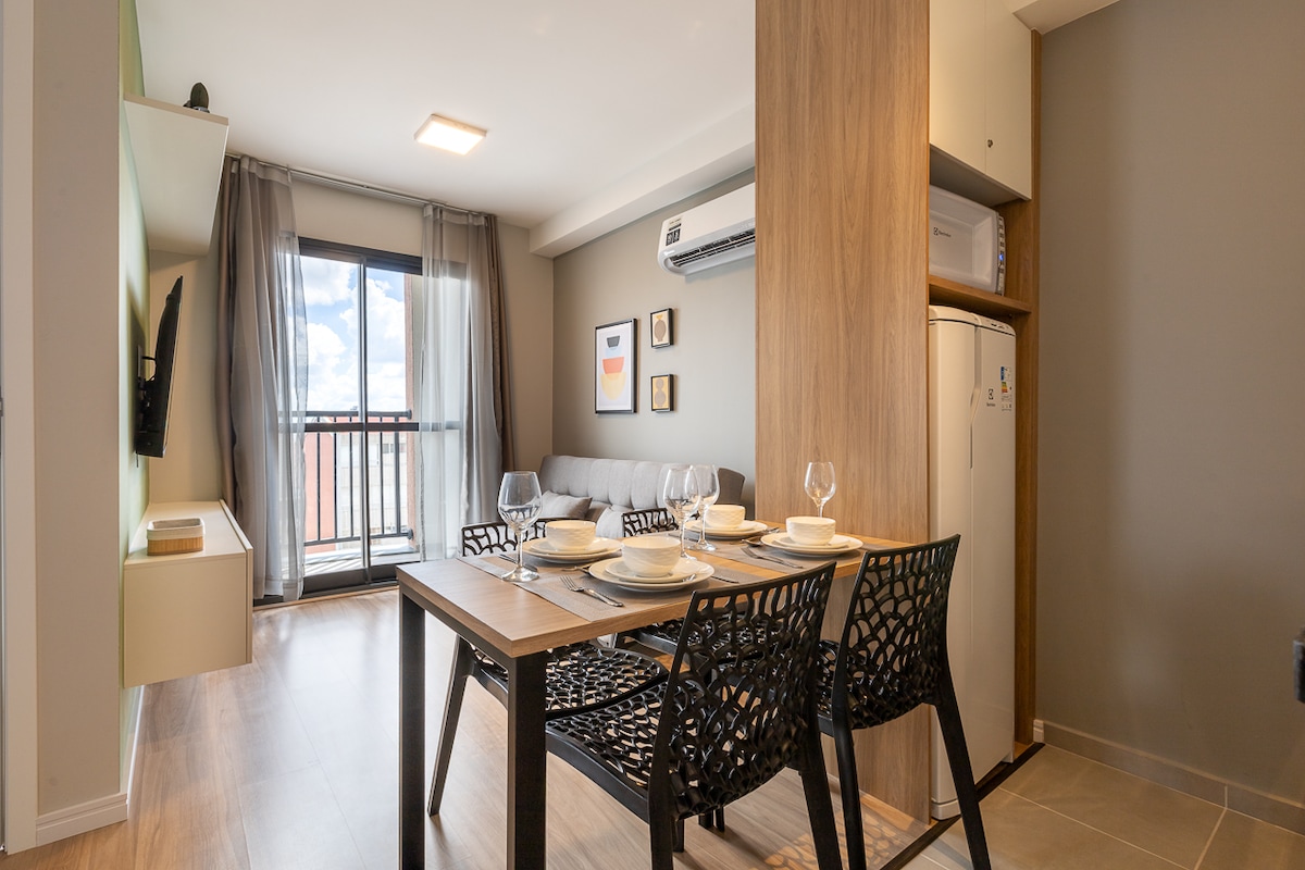 An area is shown with a dining table set for four and two stylish black chairs. A comfortable sofa is visible in the background, along with a large window that offers natural light and views. A small kitchenette is partially visible, highlighting modern appliances.