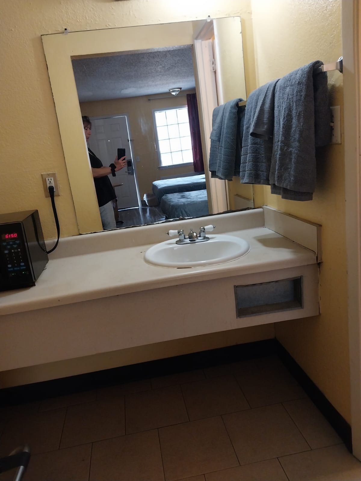 The bathroom features a simple countertop with a round sink and modern faucet. Two neatly arranged towels hang on a rack. A microwave is positioned on the counter. In the background, a glimpse of the guest room is visible, with a window allowing natural light to enter.
