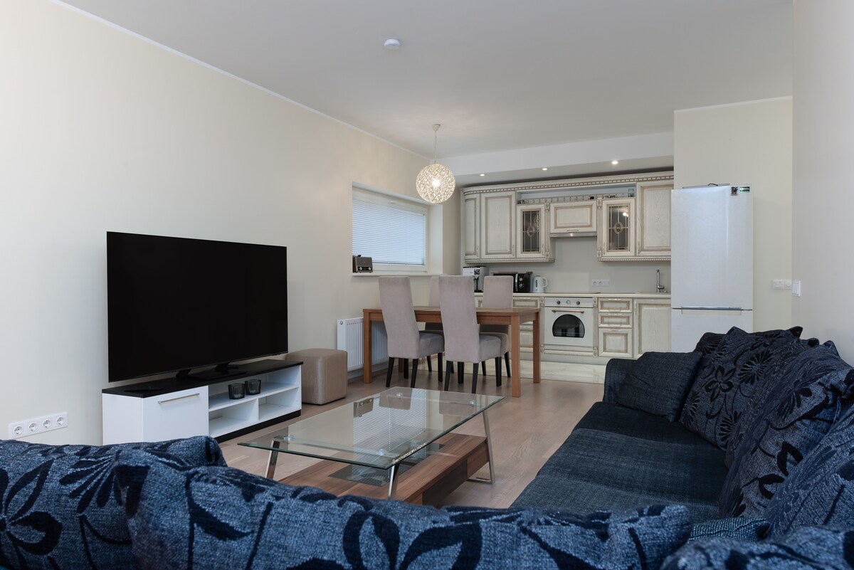 A modern living area features a large flat-screen TV opposite a spacious dark blue sofa. A glass coffee table is positioned in front, while a dining table with six chairs and a fully equipped kitchen are visible in the background.