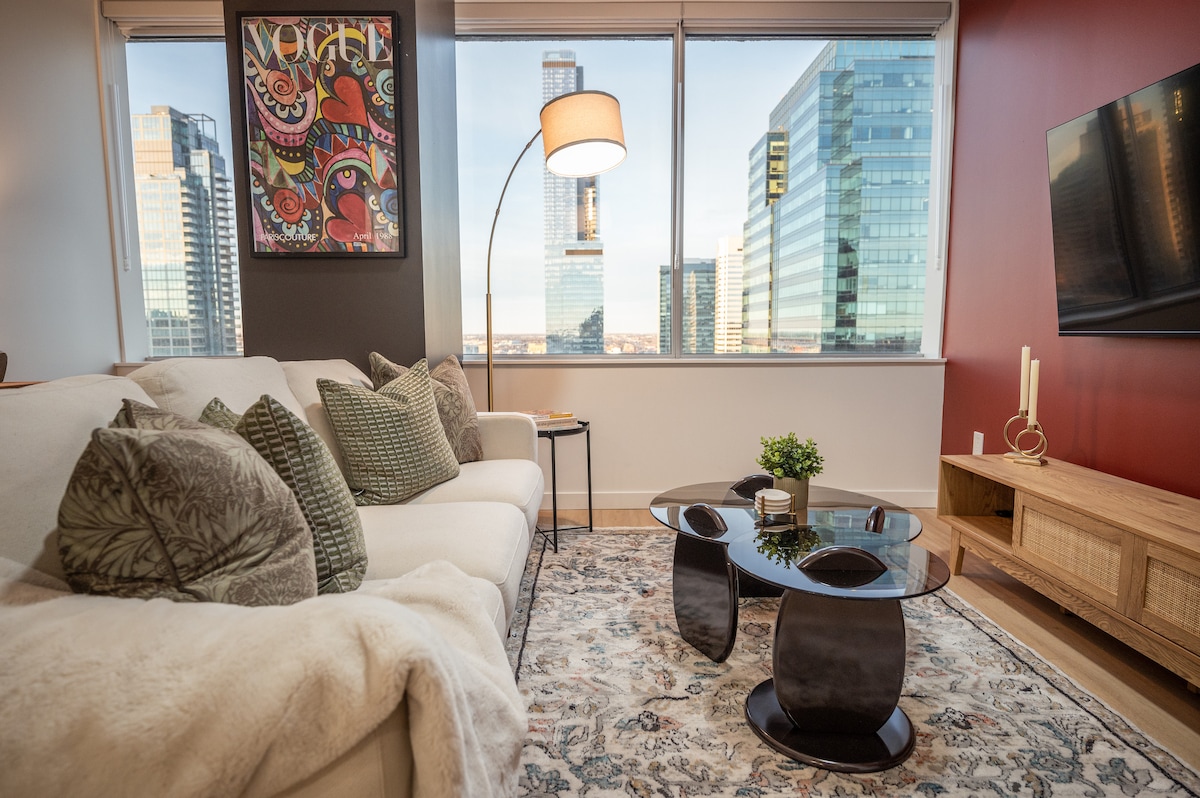 A comfortable living area features a plush white sofa adorned with various patterned cushions. A sleek glass coffee table sits in front of the sofa, complemented by a decorative plant. Large windows provide stunning views of the downtown skyline, enhancing the space's natural light.