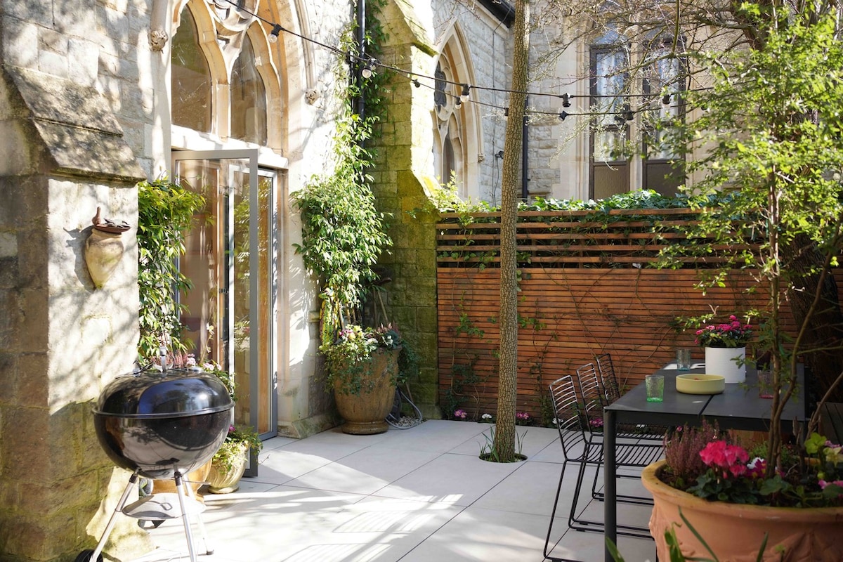 A private patio garden is bordered by heritage stone walls, showcasing a charcoal barbecue and a dining table surrounded by planters filled with vibrant flowers. Soft lighting strings are draped overhead, creating a pleasant outdoor space for dining and relaxation, framed by lush greenery.