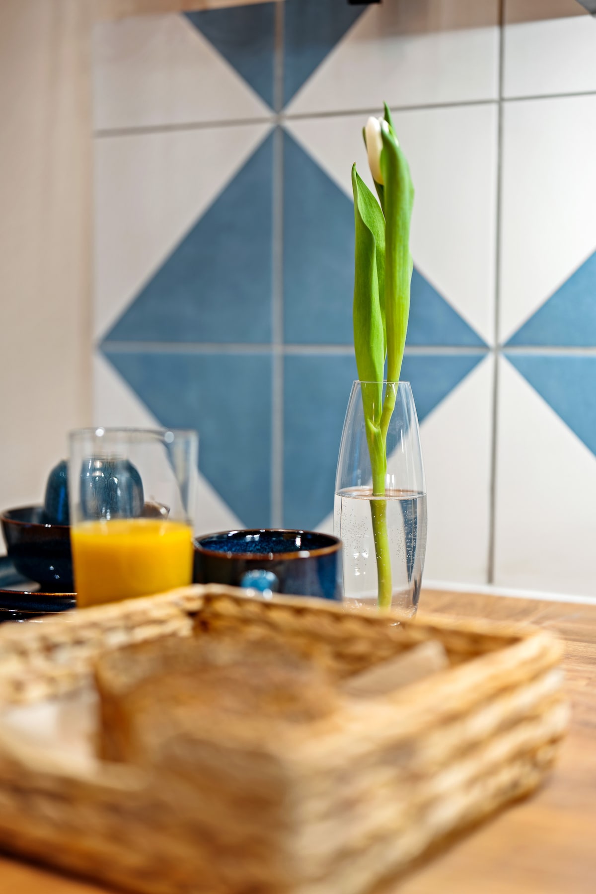 A clear glass vase holds a single green flower, placed on a wooden table. In the background, ceramic bowls and a glass with orange juice are visible, along with a woven basket containing sliced bread. Decorative blue tiles enhance the setting.