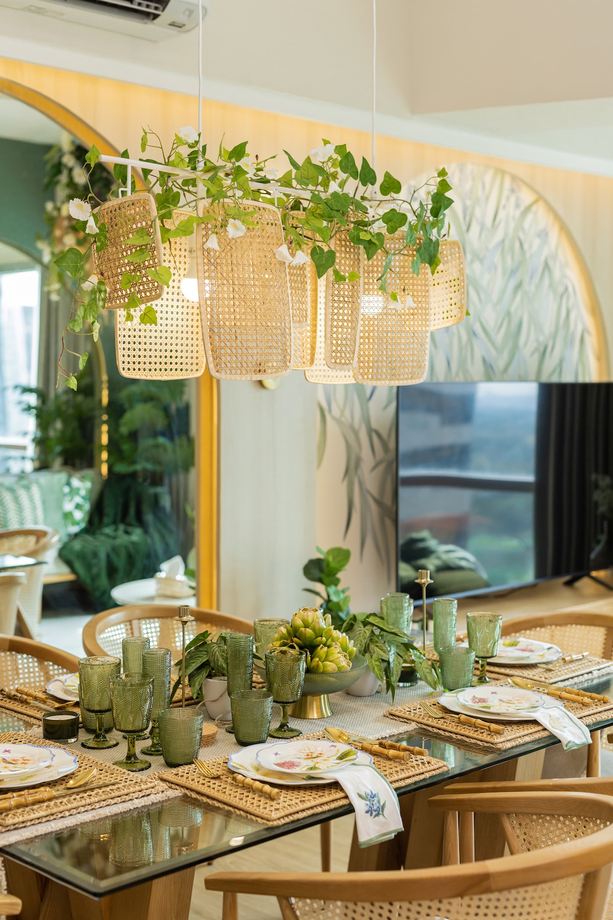 An elegant dining space features a glass table set with green tableware and clear glasses. A woven light fixture hangs above, adorned with greenery. A large television stands in the background, complementing the stylish decor, while natural light softly fills the room.