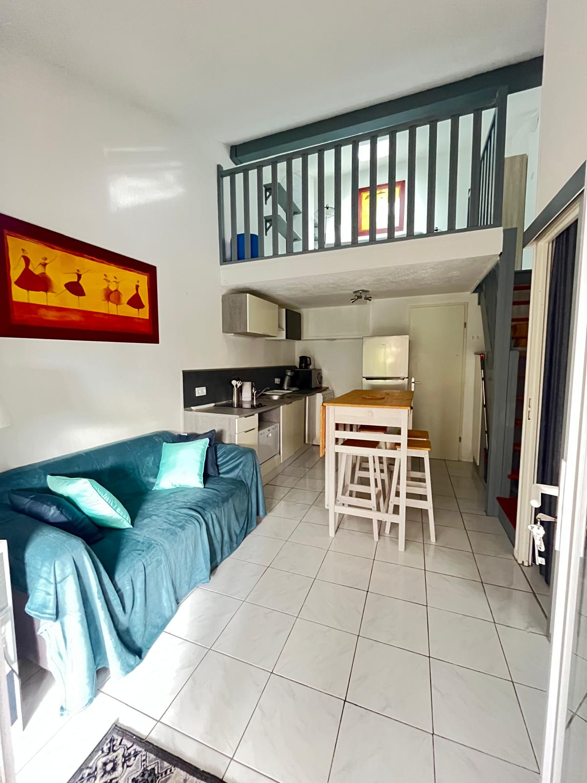 A compact living area is visible, featuring a teal sofa adorned with two green cushions. A dining space with white chairs stands adjacent to a modern kitchen equipped with a refrigerator and essential appliances. A lofted area above offers additional storage space, illuminated by natural light.